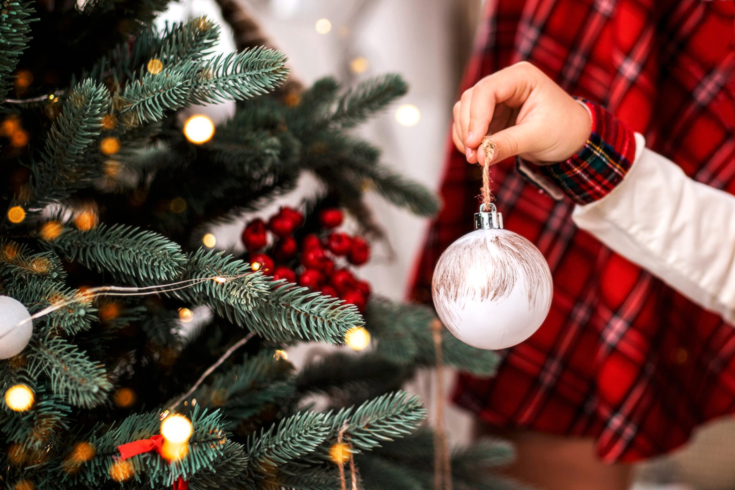 Little child's hand decorating Christmas tree white ball indoors. Close up.