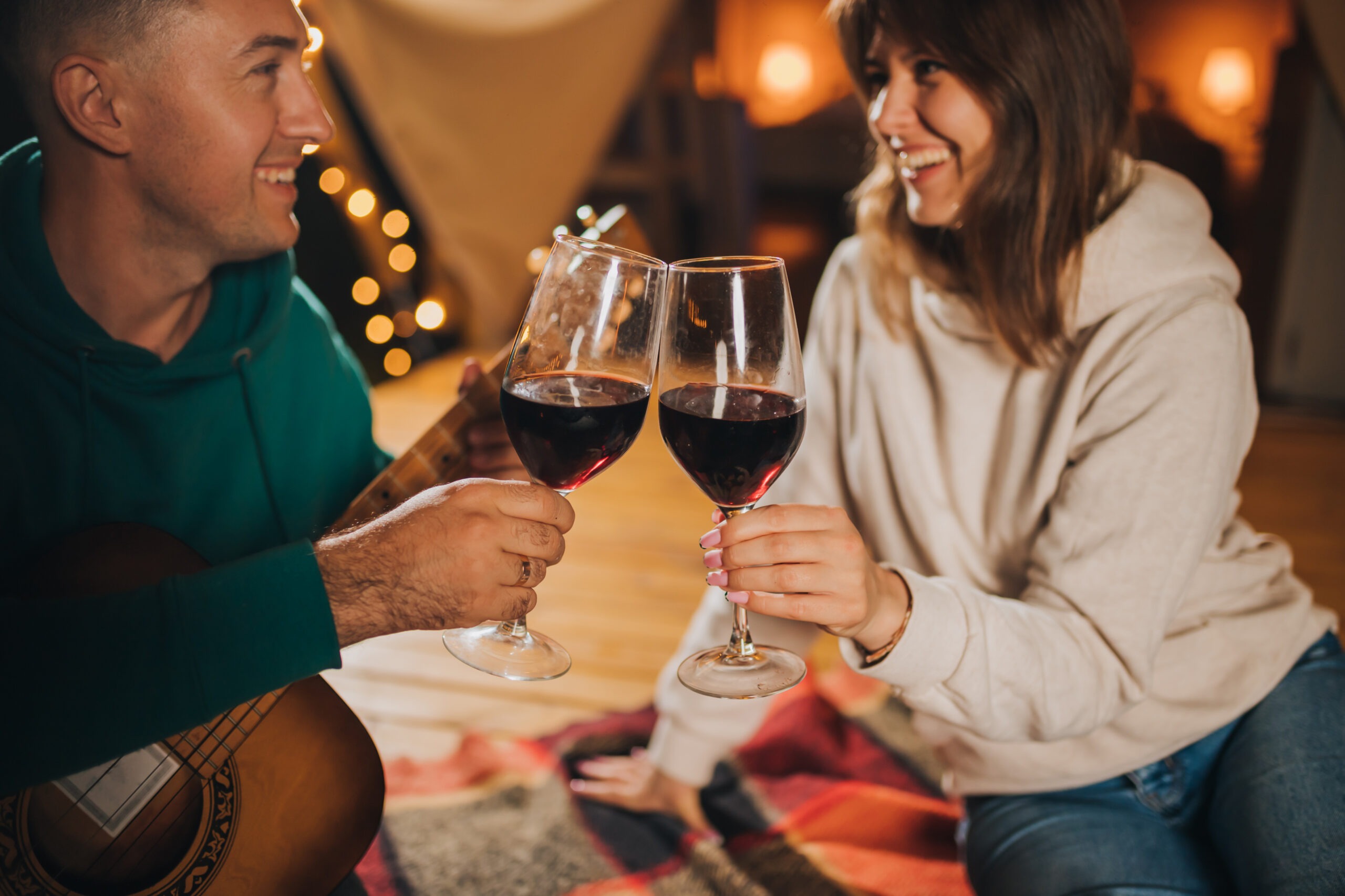 Happy couple relaxing in glamping on autumn evening, drinking wine and playing guitar near cozy bonfire. Luxury camping tent for outdoor recreation and recreation. Lifestyle concept